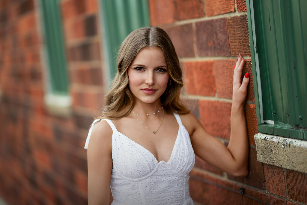 A high school senior in a white dress leans on a brick wall before her grand view university graduation