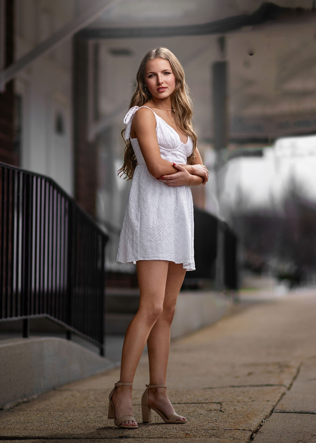 A high school senior in a white dress stands with arms crossed in an alley after visiting hair salons in pella, iowa