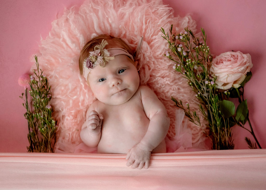 A newborn baby lays under a pink blanket with flowers and a pink headband with eyes open