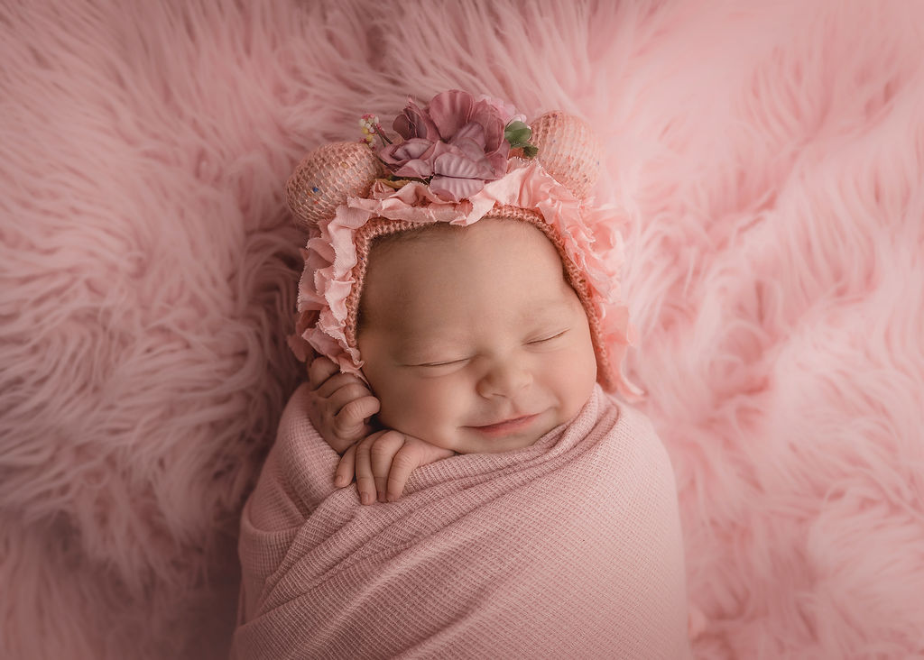 A newborn baby girl giggles in her sleep in a pink floral headpiece and pink swaddle on a matching fur blanket after meeting iowa baby lady