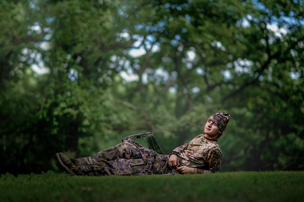 A knoxville high school iowa senior hunter lays in a lawn in camo with his hunting bow