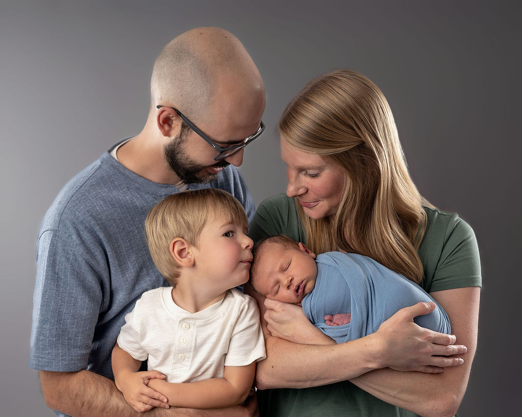 Happy parents stand in a studio holding their newborn baby and toddler son in their arms