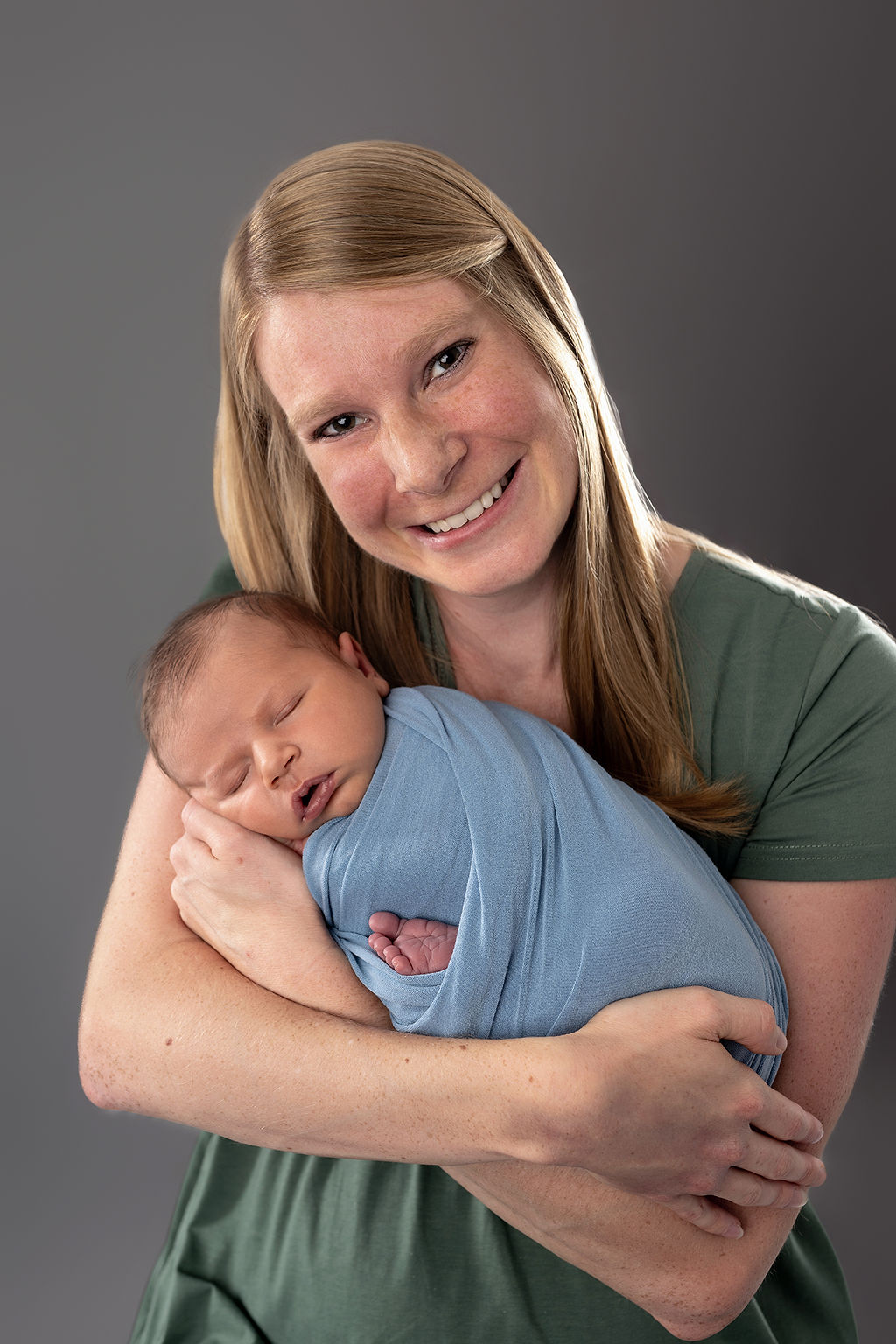 A smiling new mother in a green dress cradles her sleeping newborn baby in her arms in a blue swaddle in a studio after meeting a lactation consultant in Des Moines