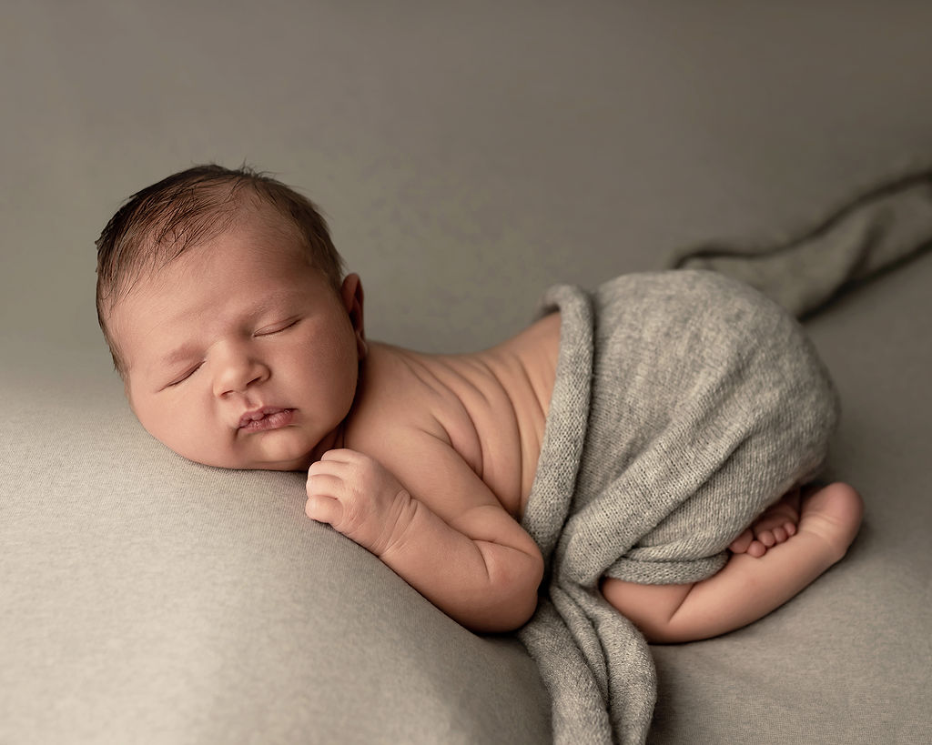 A sleeping newborn baby in froggy pose under a grey blanket in a studio after visiting Little Wonders Wellness