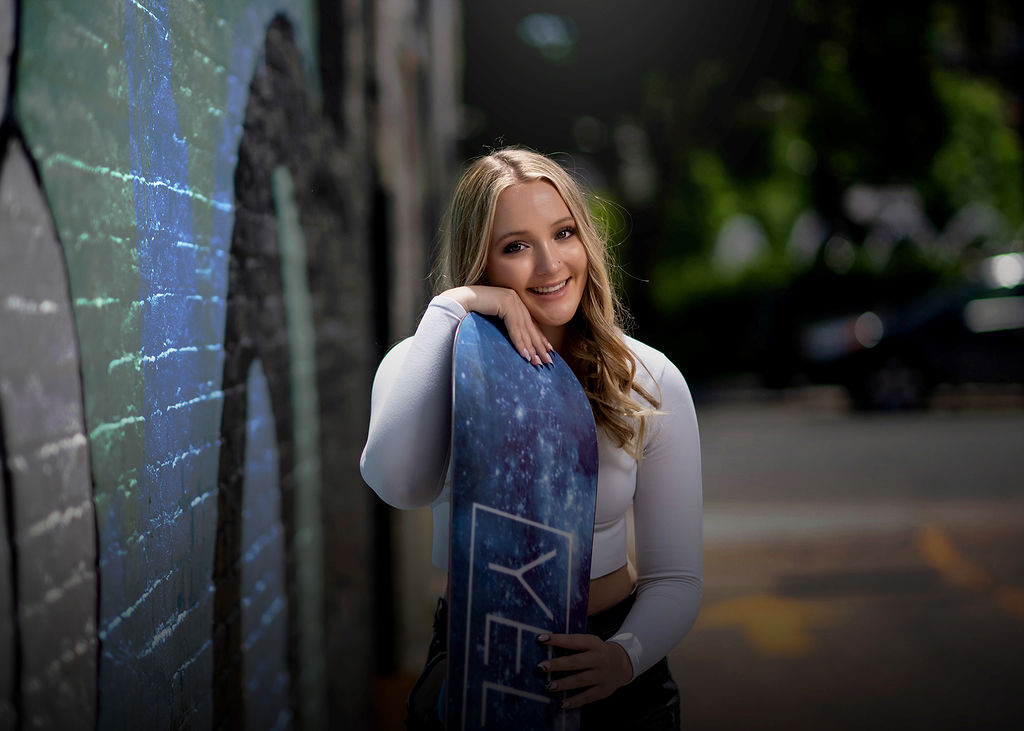 A happy blonde girl leans on a snowboard smiling in a white shirt in an alley next to graffiti