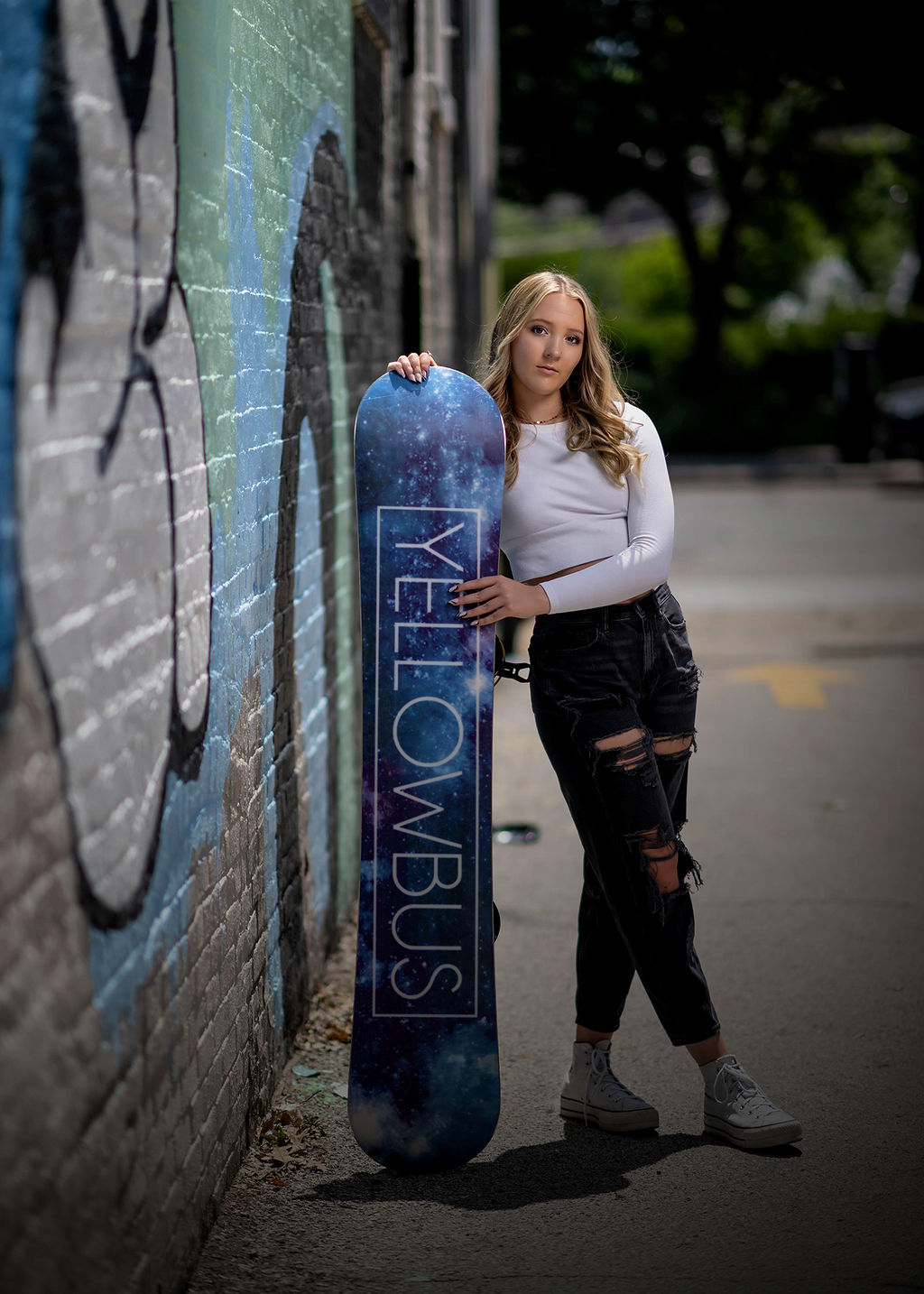 A high school senior leans on a snowboard in an alley in a white top and ripped jeans after visiting a nail salon in pella, iowa