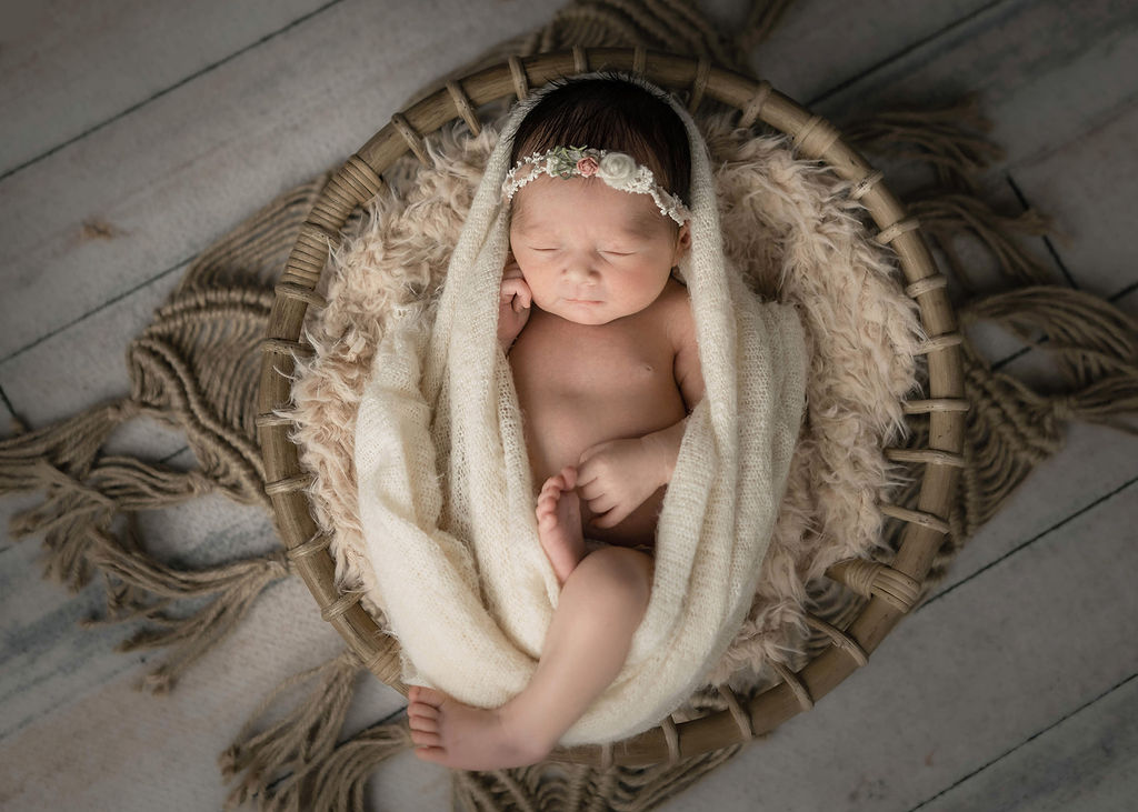 A newborn baby sleeps with a floral headband in a wicker basket on a wooden floor