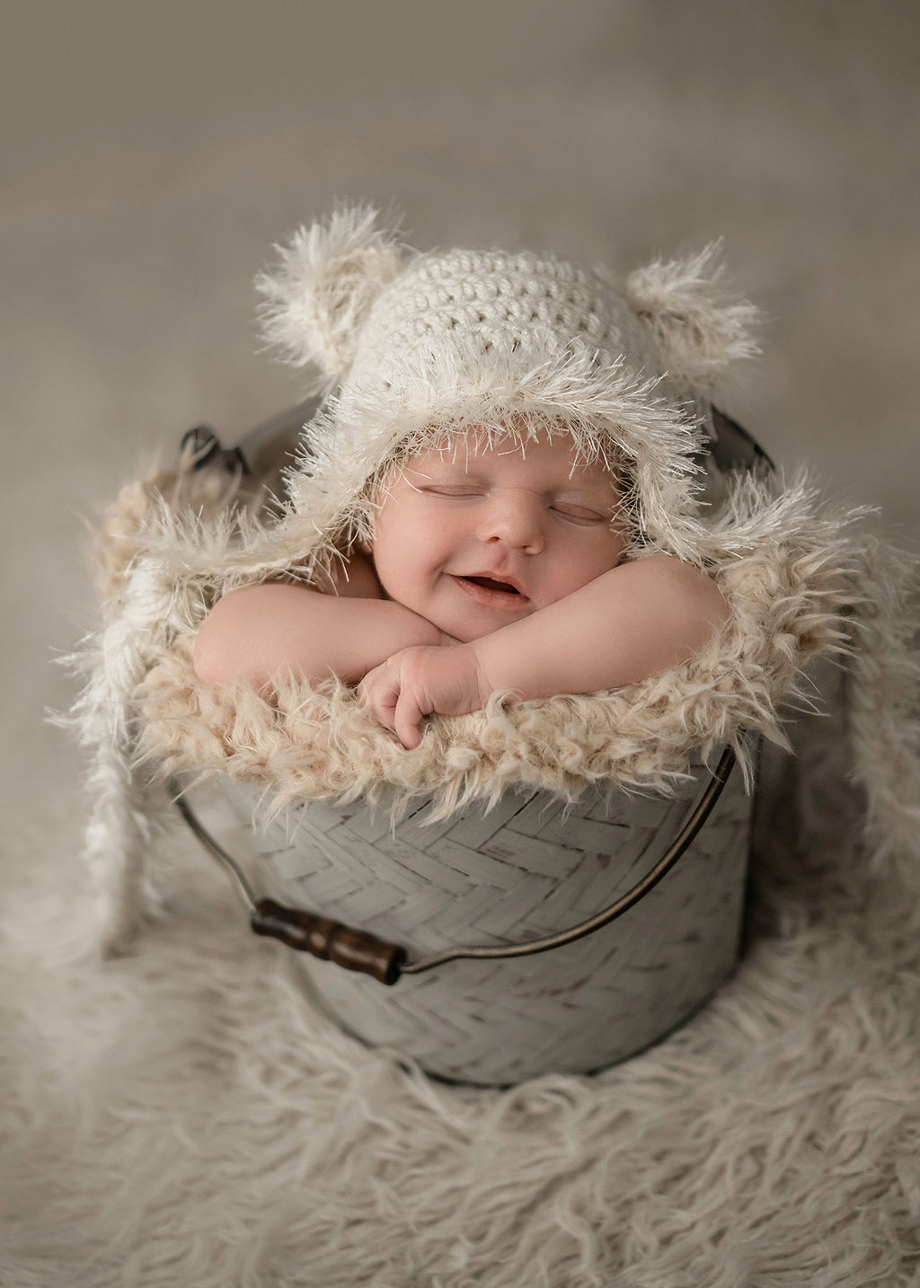 A newborn baby smiles in its sleeps in a metal bucket and knit bear hat after meeting nannies in des moines