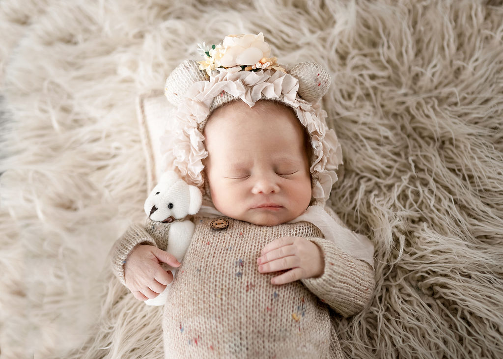 A newborn bay sleeps in tan knit onesie on a matching fur blanket with a white felt teddy after meeting nannies in des moines