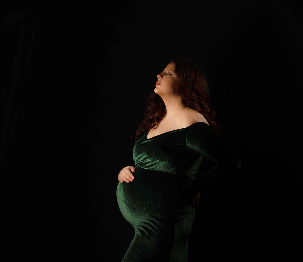 A mother to be looks up with a hand on her bump in a green velvet maternity gown