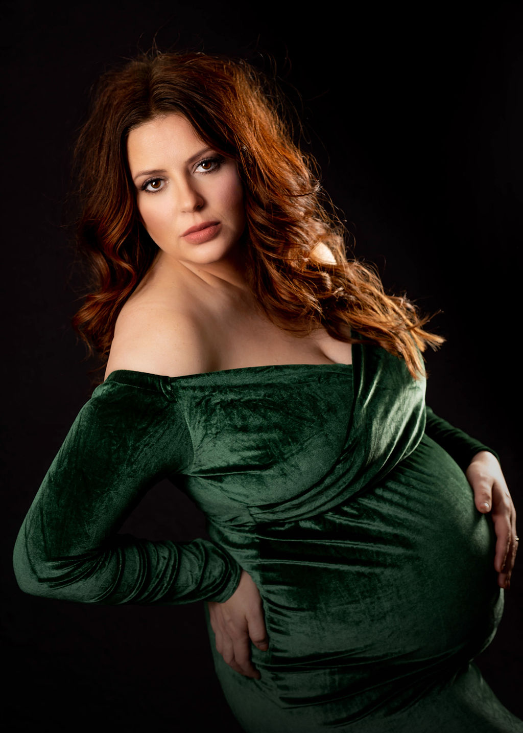 A mother to be stands in a studio with a hand on her bump in a green velvet maternity gown after visiting an obgyn in des moines