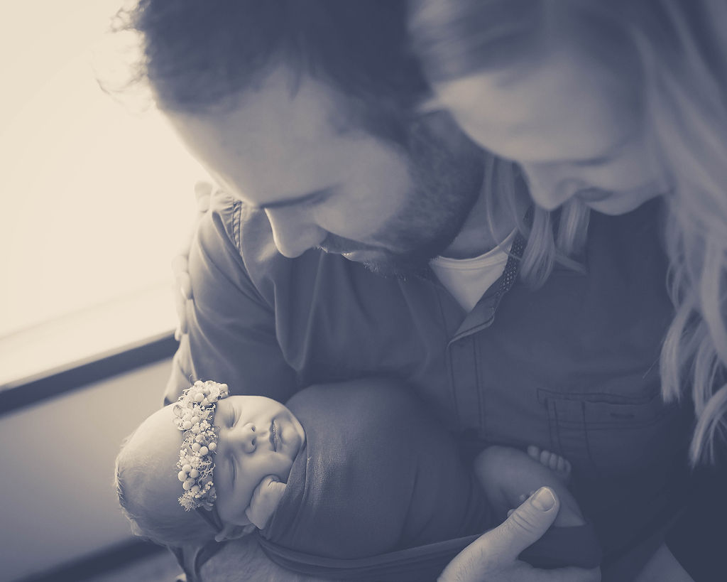 A newborn baby sleeps in dad's arms as mom looks over his shoulder in a window