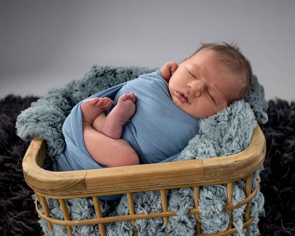 A newborn baby sleeps in a blue onesie in a wooden basket in a studio