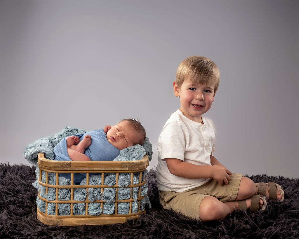 A happy toddler in a white shirt sits with his newborn baby brother sleeping in a wooden basket in a studio after a pediatric chiropractor in Des Moines