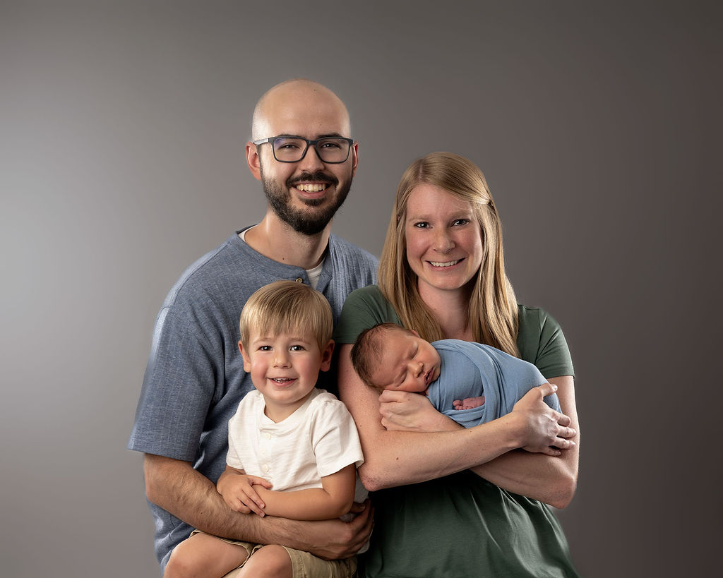 A happy mom and dad smile while sitting in a studio with their newborn and toddler son in their laps after visiting a pediatric chiropractor in Des Moines