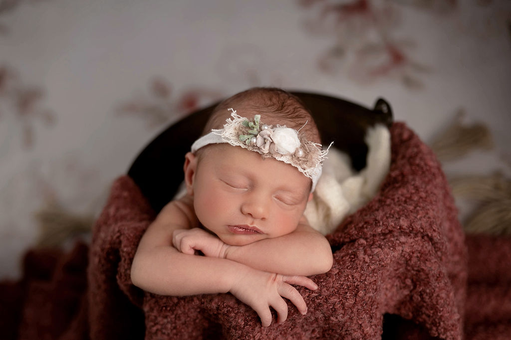 A newborn baby sleeps in a bucket wearing a white floral headband in a studio