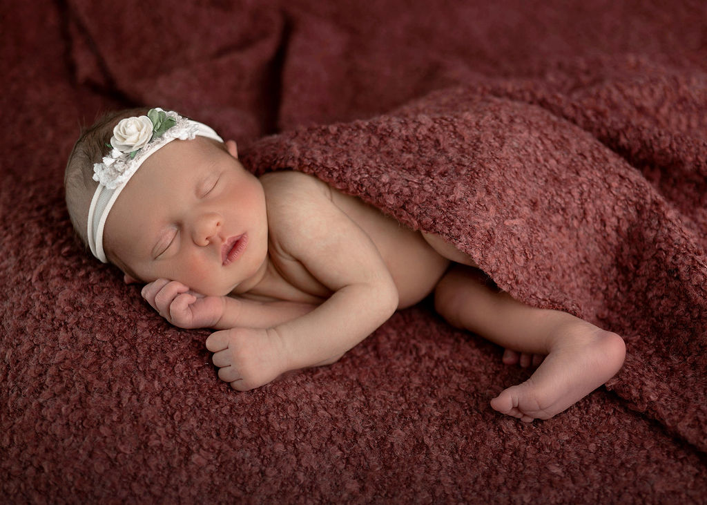 A newborn baby girl in a white floral headband sleeps under a red blanket in a studio after finding a pediatric dentist in des moines