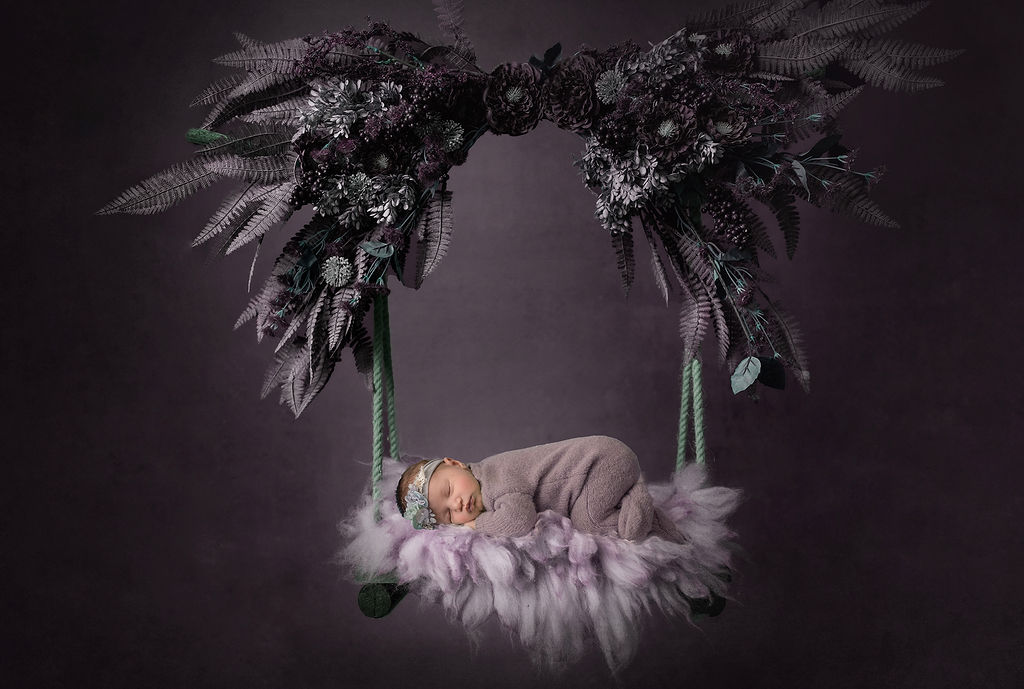 A newborn baby sleeps on a purple floral covered swing in a studio