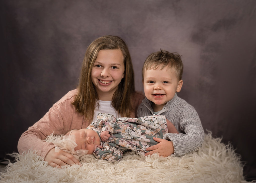 Happy young brother and sister sit behind their sleeping newborn baby sister in a studio after visiting pediatricians in des moines