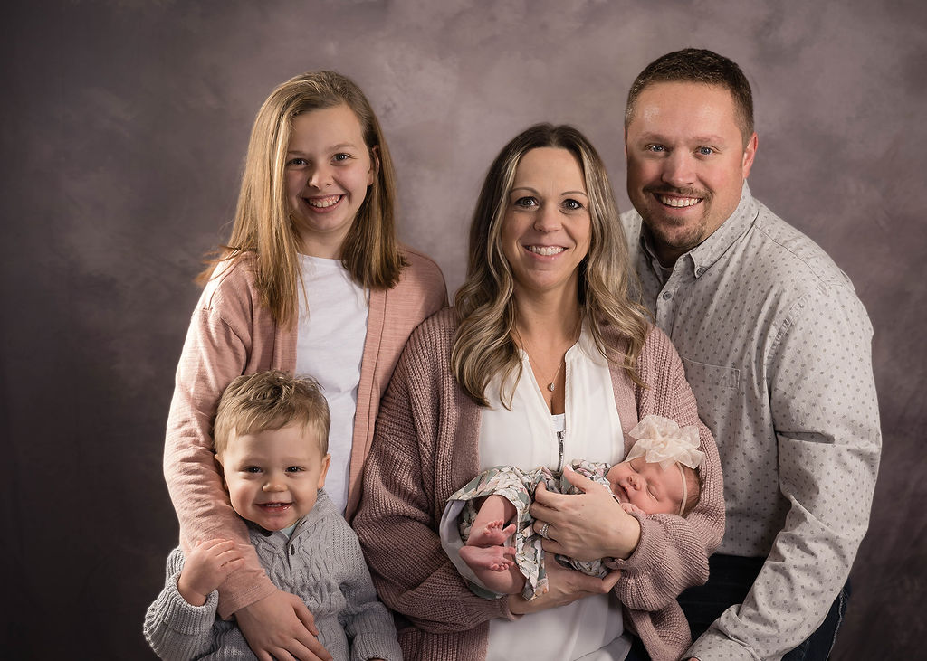 A happy mom and dad stand in a studio with their young daughter and baby son and newborn baby in mom's arms after finding pediatricians in des moines