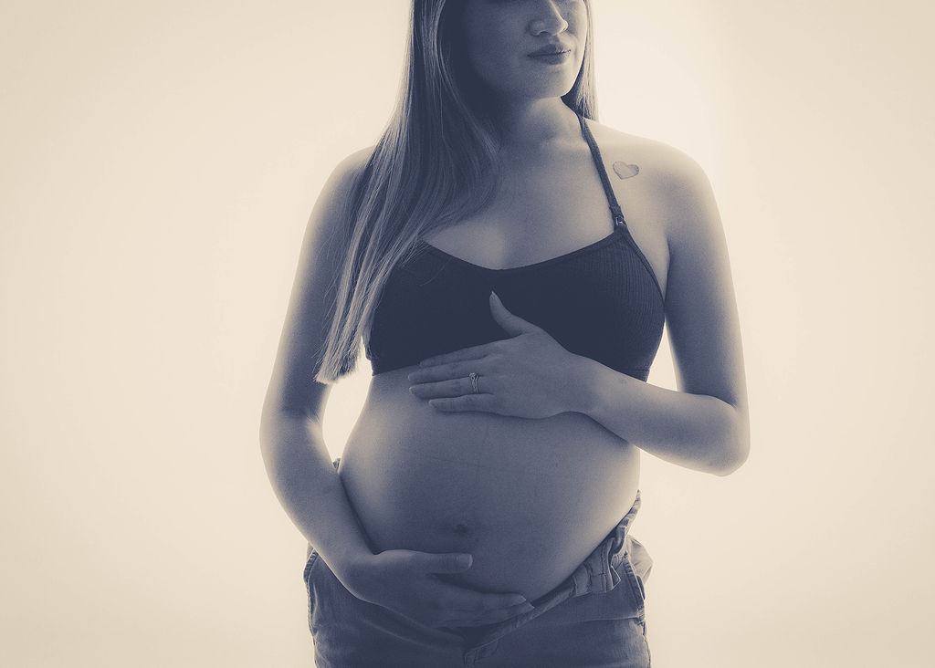 A pregnant woman holds her bump while standing in a studio in a bra and jeans