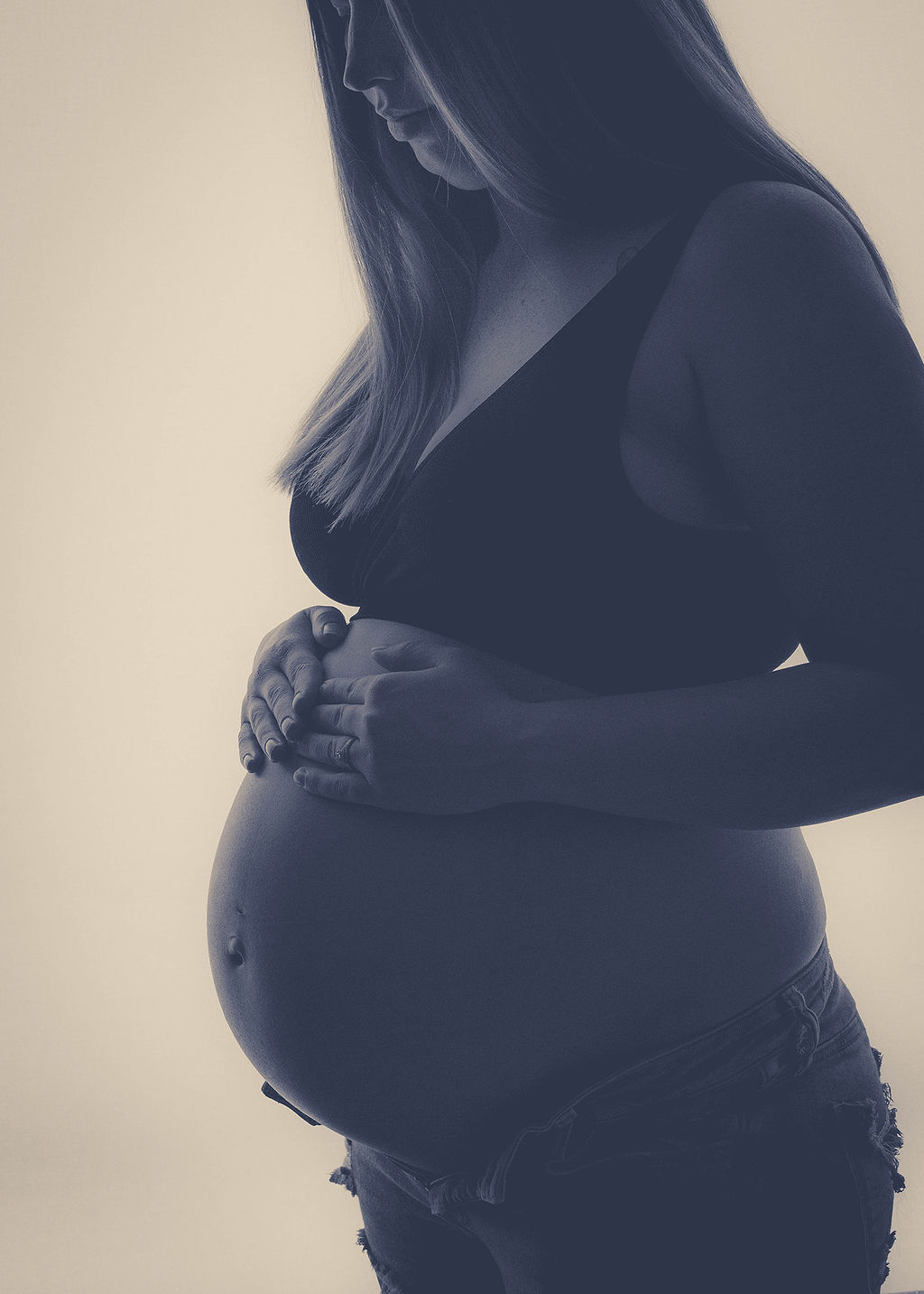 A mom to be rests her hands on her bump while standing in a studio in a sports bra and jeans after visiting a pella obgyn