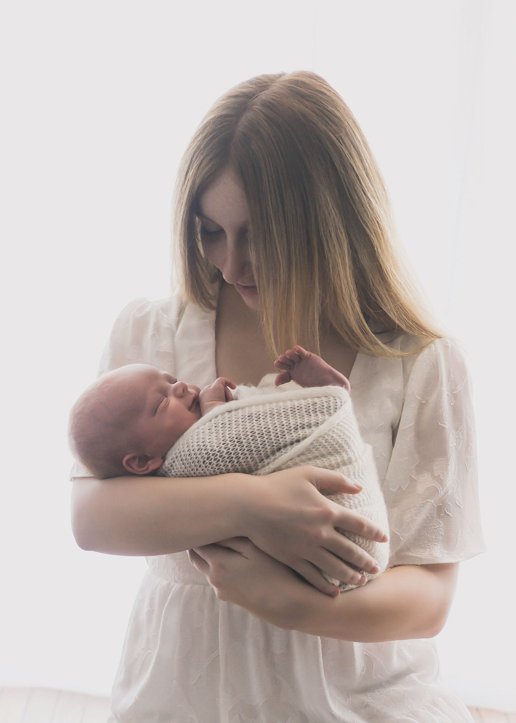 A new mom cradles her sleeping newborn baby while standing in a studio in a white dress