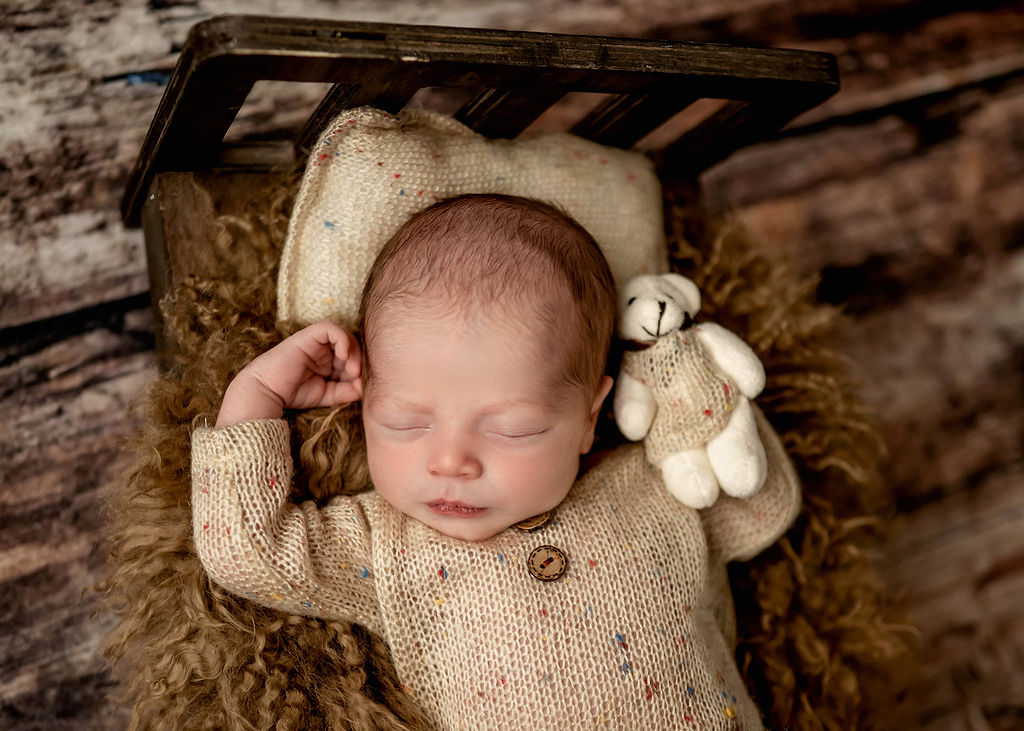 A sleeping newborn baby in a knit beige onesie in a tiny wooden bed with a tiny bear