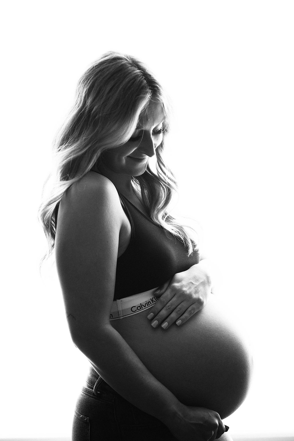 A smiling mom to be stands in a studio in a sports bra and open jeans smiling at her bump after a prenatal massage in des moines