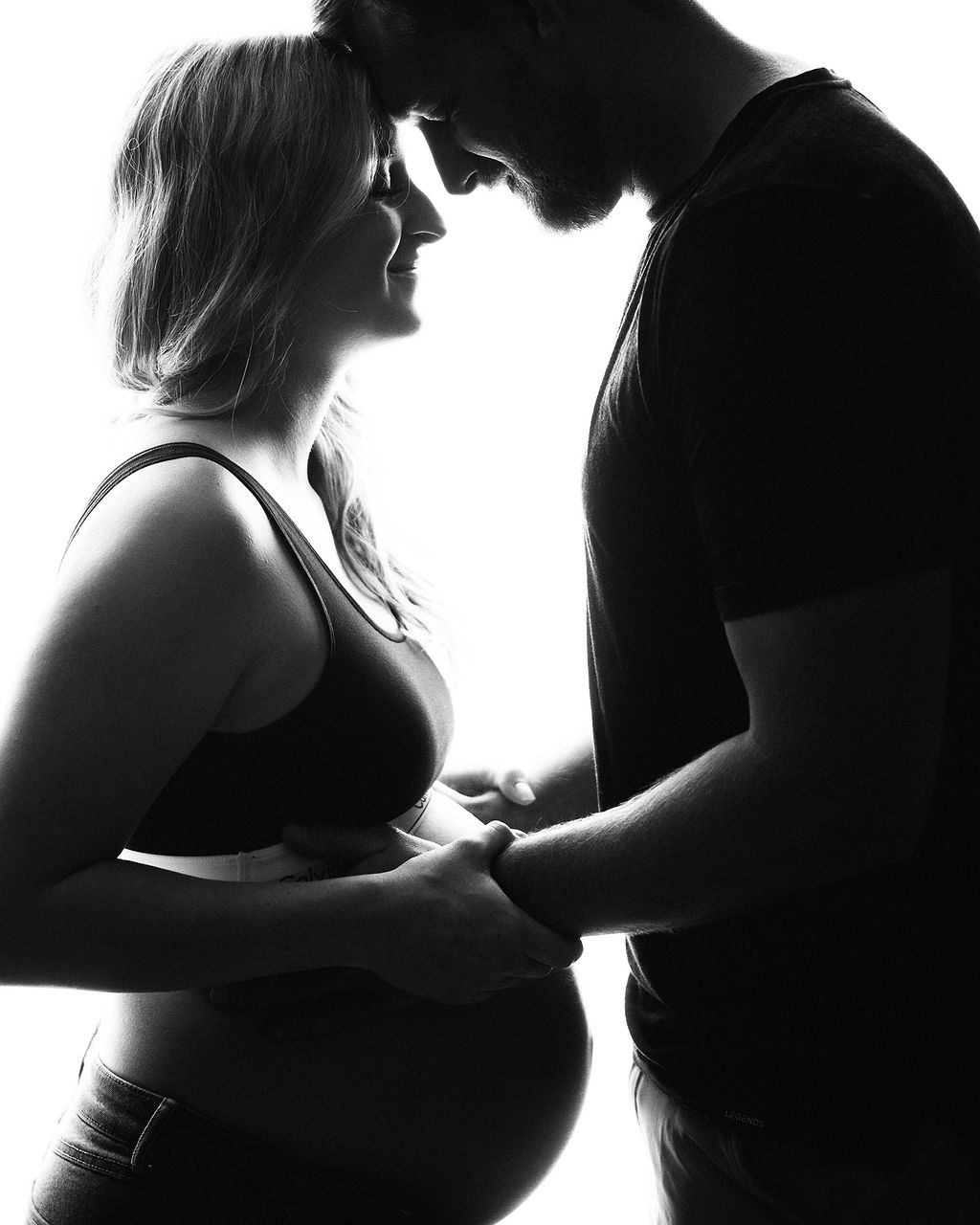 Happy expecting parents stand in silhouette with hands on the bump and foreheads touching after a prenatal massage in des moines