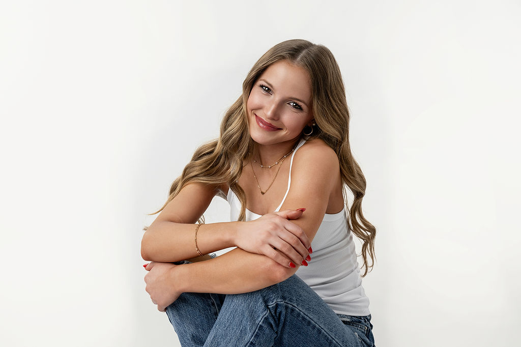 A happy high school senior sits with arms on her knees in a white studio