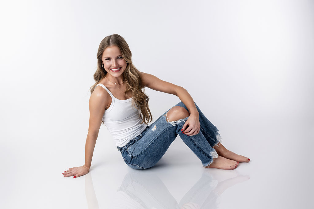 A woman sits on the floor of a studio in a white top and ripped jeans smiling big after visiting tanning salons in pella, iowa