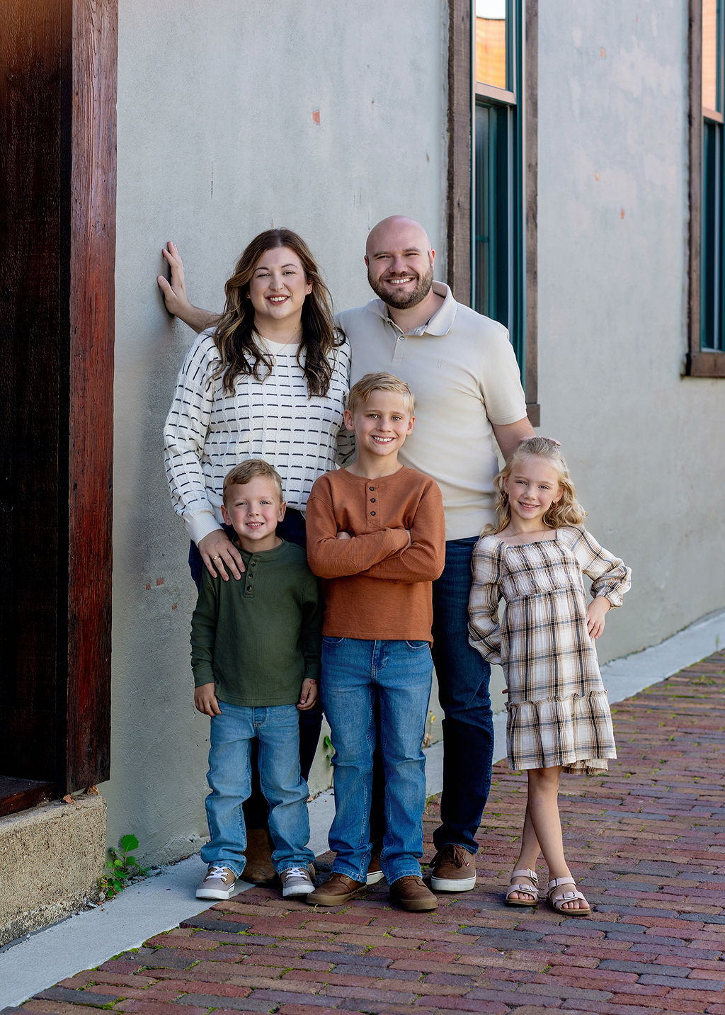 A happy family of 5 stand in a brick paved alley smiling