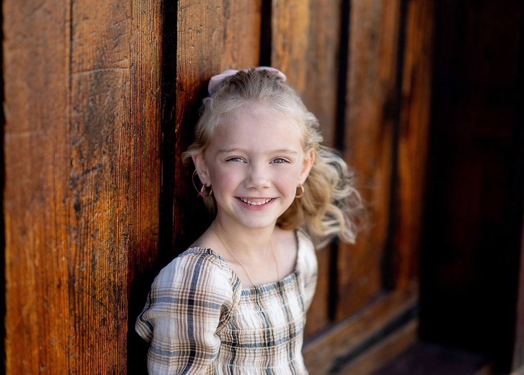 A young girl leans on a wooden wall in a plaid dress smiling after some things to do in Des Moines with kids