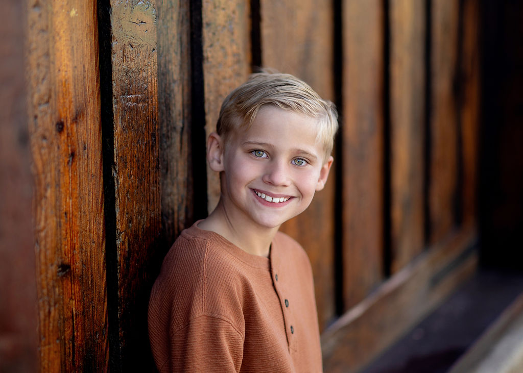 A young boy leans on a wooden wall in an orange henley smiling during things to do in Des Moines with kids