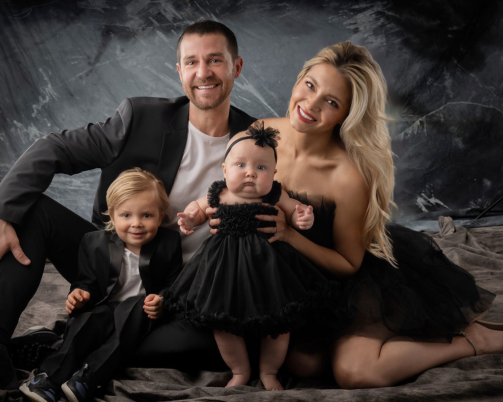 A smiling mom and dad sit on a studio floor with their toddler son and baby daughter all in black