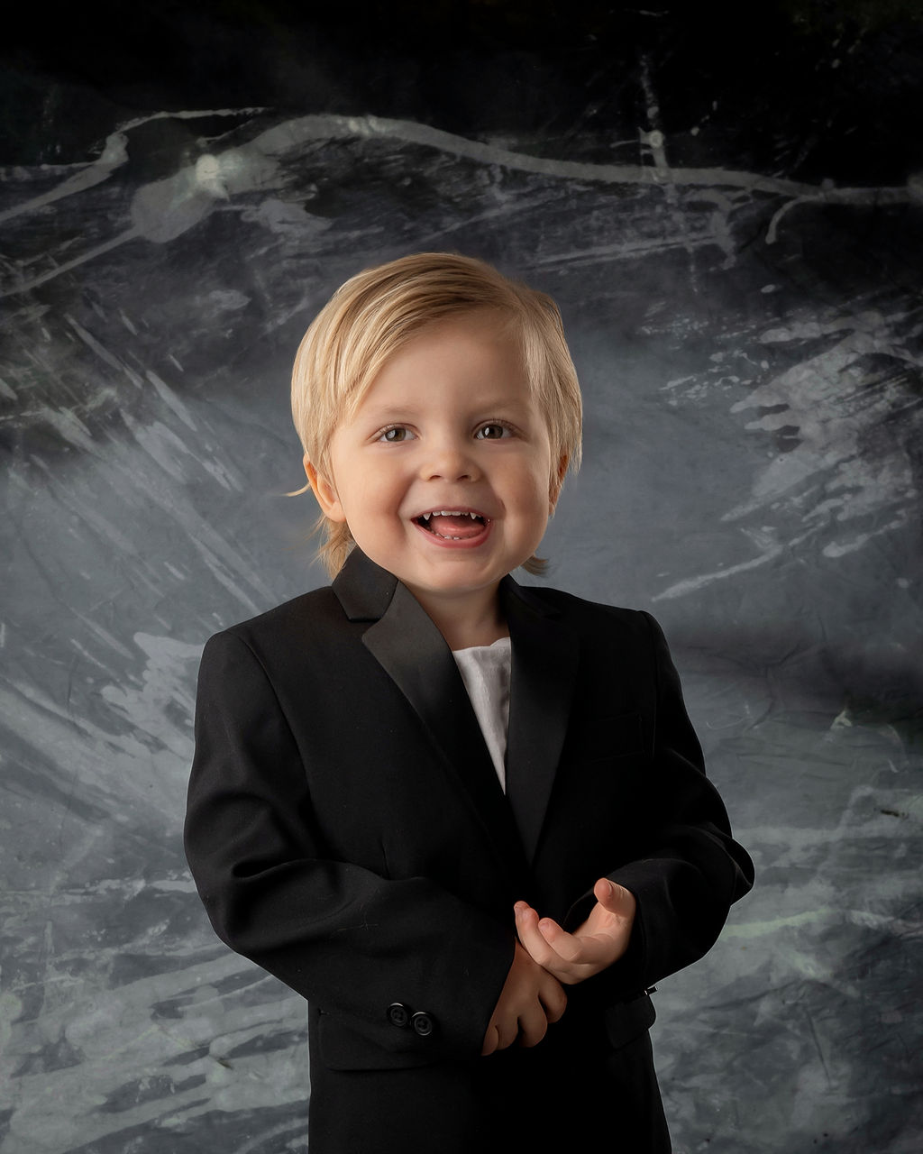 A smiling toddler boy in a black suit jacket stands in a studio after some things to do in Des Moines with toddlers