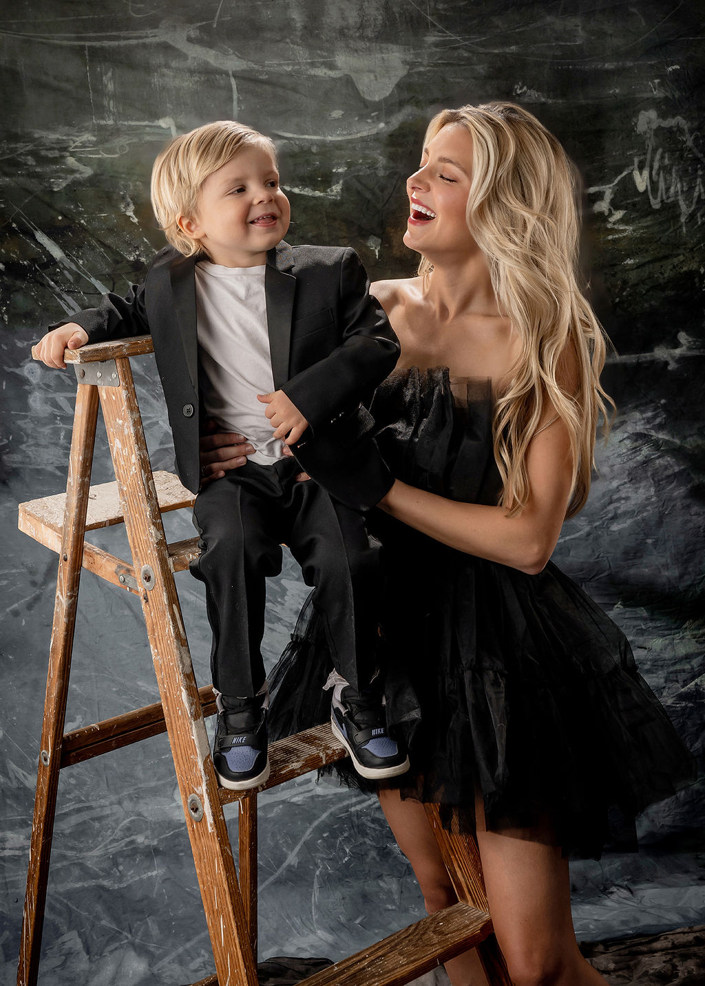 A happy toddler sits on a wooden ladder in a studio with happy mom holding him during things to do in Des Moines with toddlers