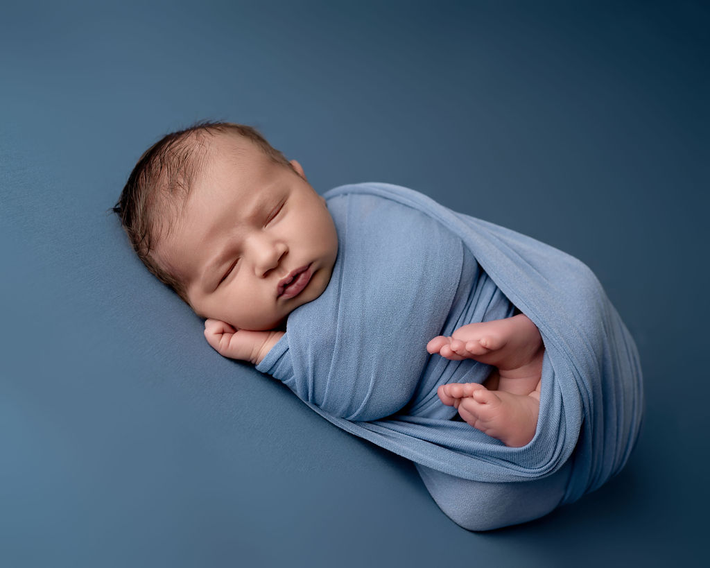 A newborn baby sleeps on its side in a blue swaddle with feet sticking out