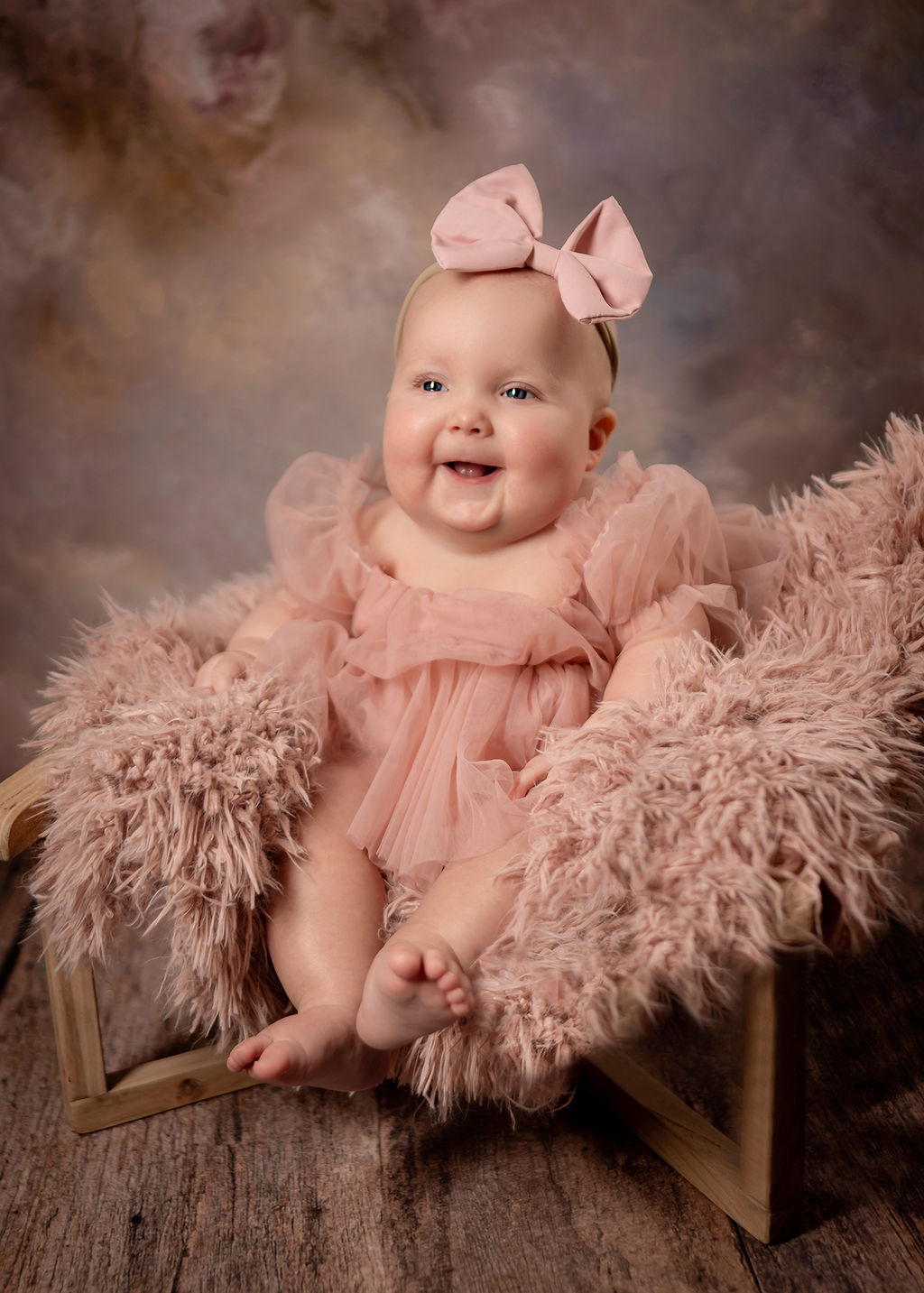 A smiling baby in a pink dress sits in a shag bench in a studio