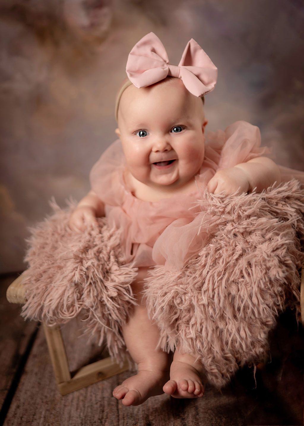 A smiling baby in a pink dress sits on a tiny bench in a studio with a pink bow before visiting a Daycare in Pella, Iowa