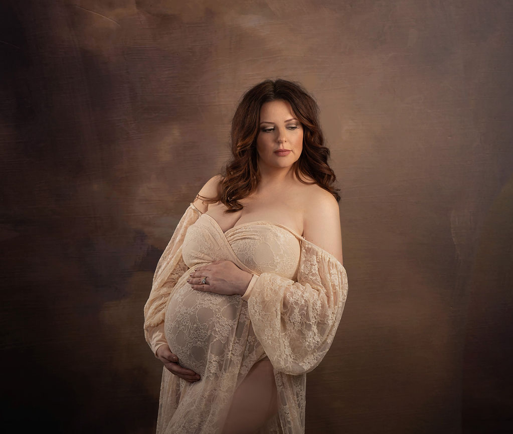 A mom to be in a lace dress gazes down her shoulder while standing in a studio