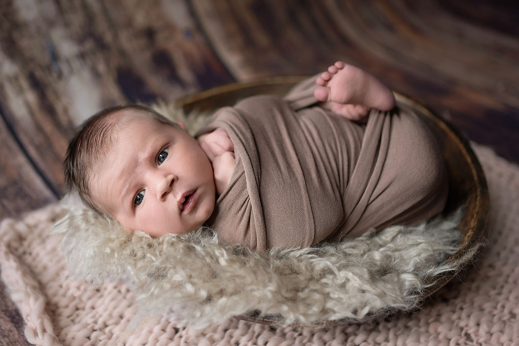 A newborn lays in a wooden bowl in a brown swaddle with eyes open after visiting Ivory Soul