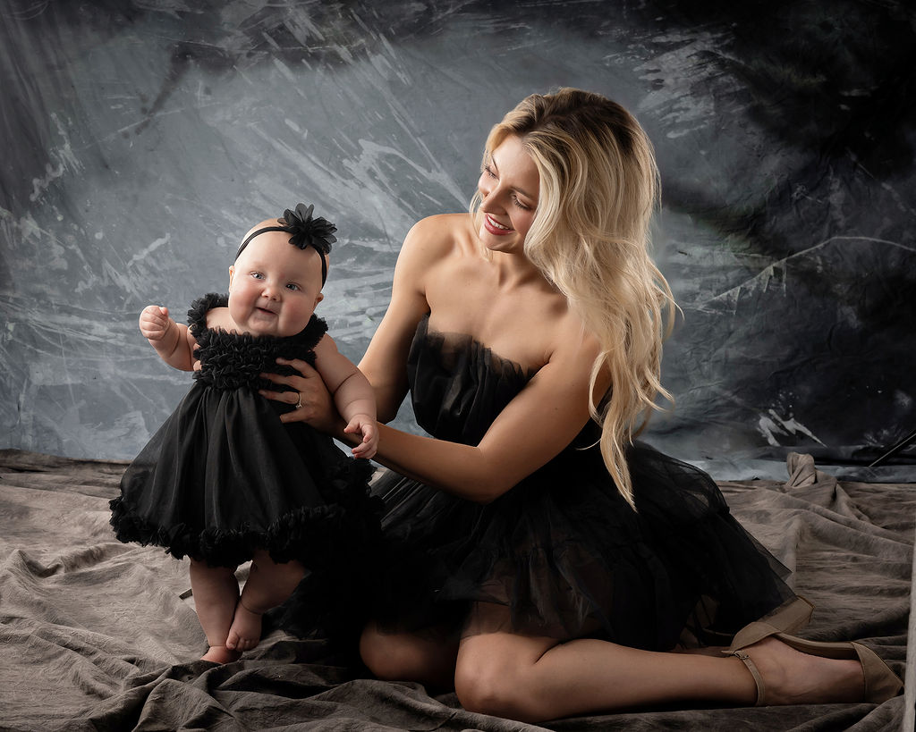 A mother in a black dress sits on the floor of a studio helping her baby daughter in a matching dress stand after visiting Kinetic Edge Physical Therapy