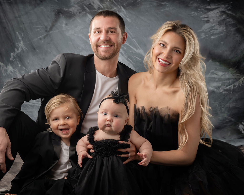 A happy family with two babies sit in black in a studio smiling after visiting Kinetic Edge Physical Therapy