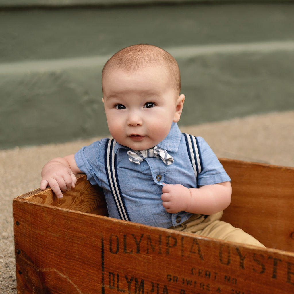 A baby in a blue shirt and overalls sits in a wooden apple box after visiting Little Children's Boutique