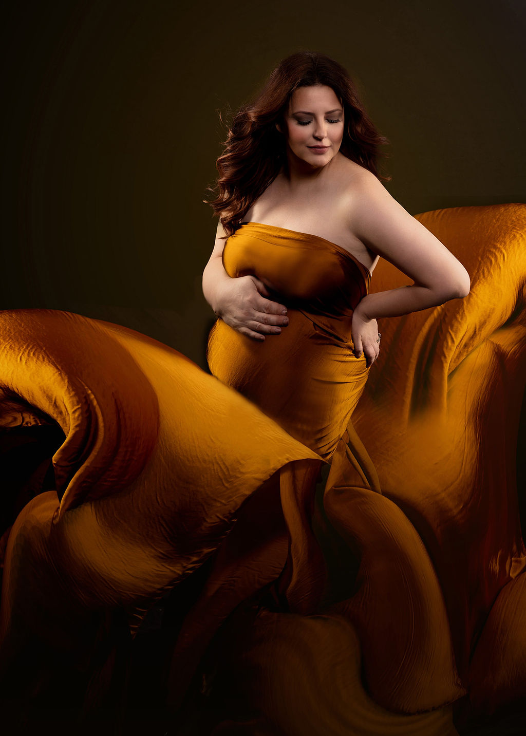 A pregnant woman stands in a studio wrapped in a gold sheet that is flowing all around her