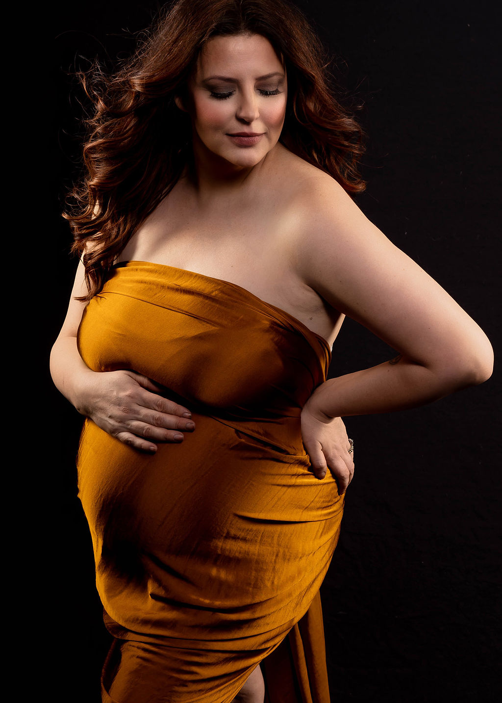 A mom to be wrapped in a gold sheet stands in a studio with a hand on her hip after visiting mercyone birth center