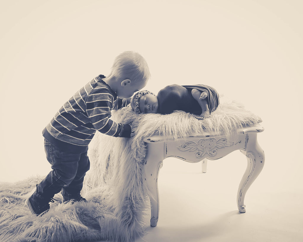 A toddler leans over his sleeping newborn sister on a bench in a studio after a parenting class in pella, iowa