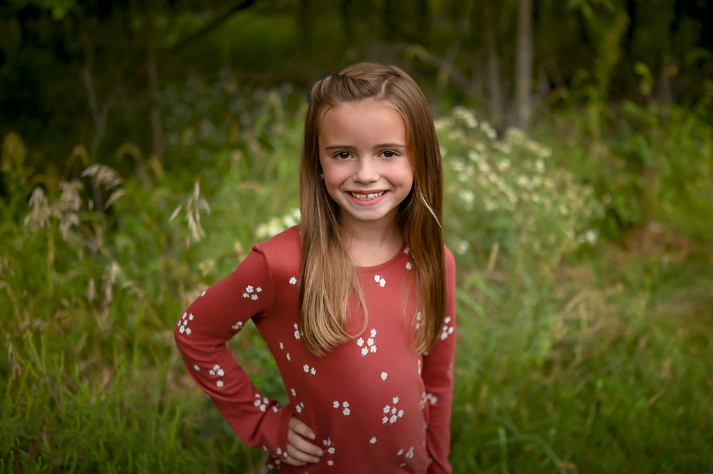 A young girl in a red flower dress with a hand on her hip smiling in a forest after visiting Pella Family Dentistry